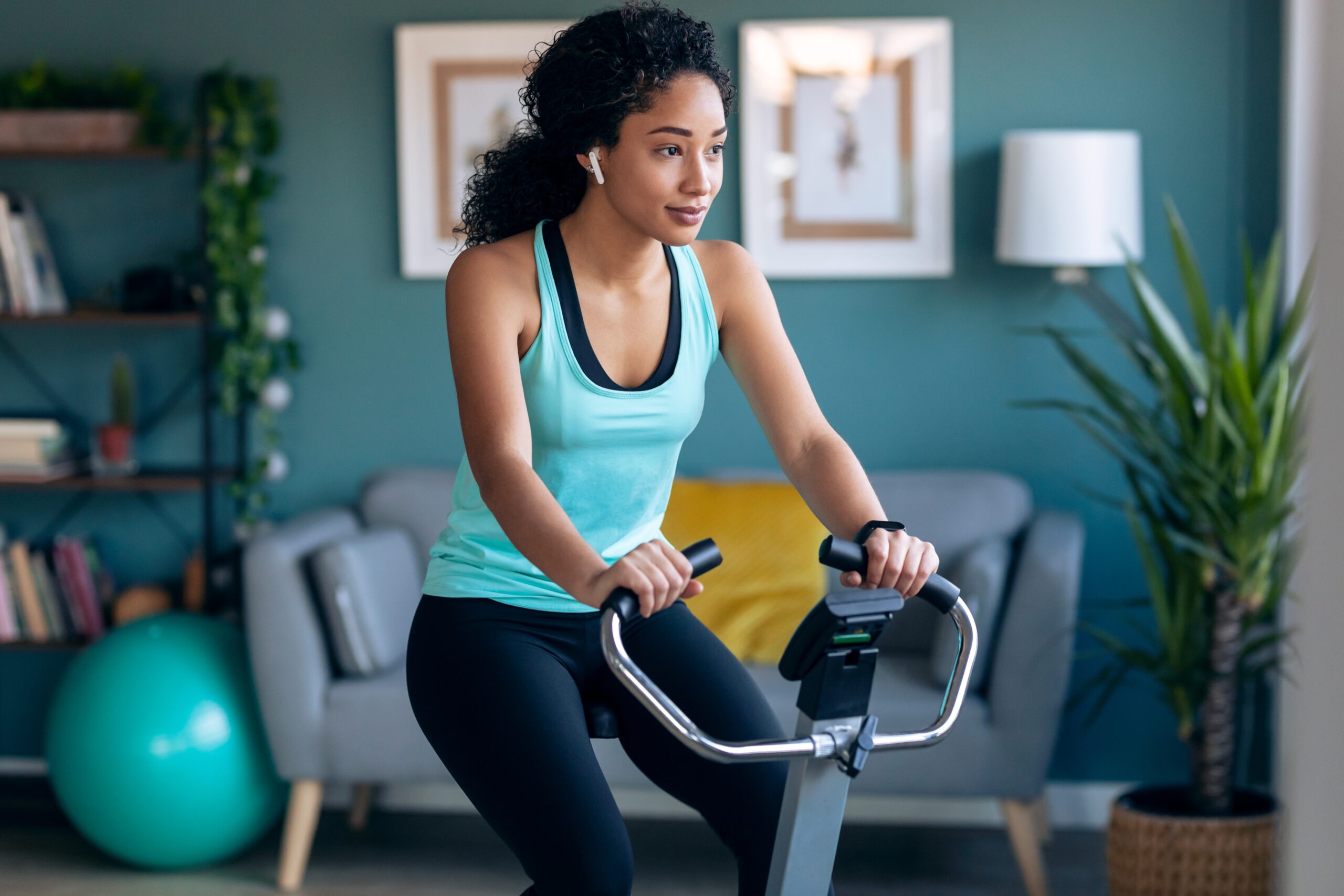 Woman in athletic wear cycling indoors on an exercise bike, focused on her workout. A bright, modern home gym setting is visible in the background.