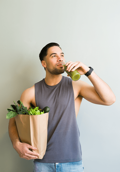 Man in a sleeveless shirt holds a bag of fresh greens and drinks a green smoothie, promoting a healthy lifestyle and nutrition.